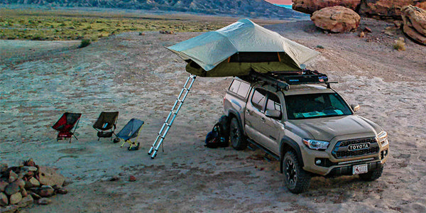 A truck with a rooftop tent is set up in the desert. There are folding camp chairs and a rock ring to the left.
