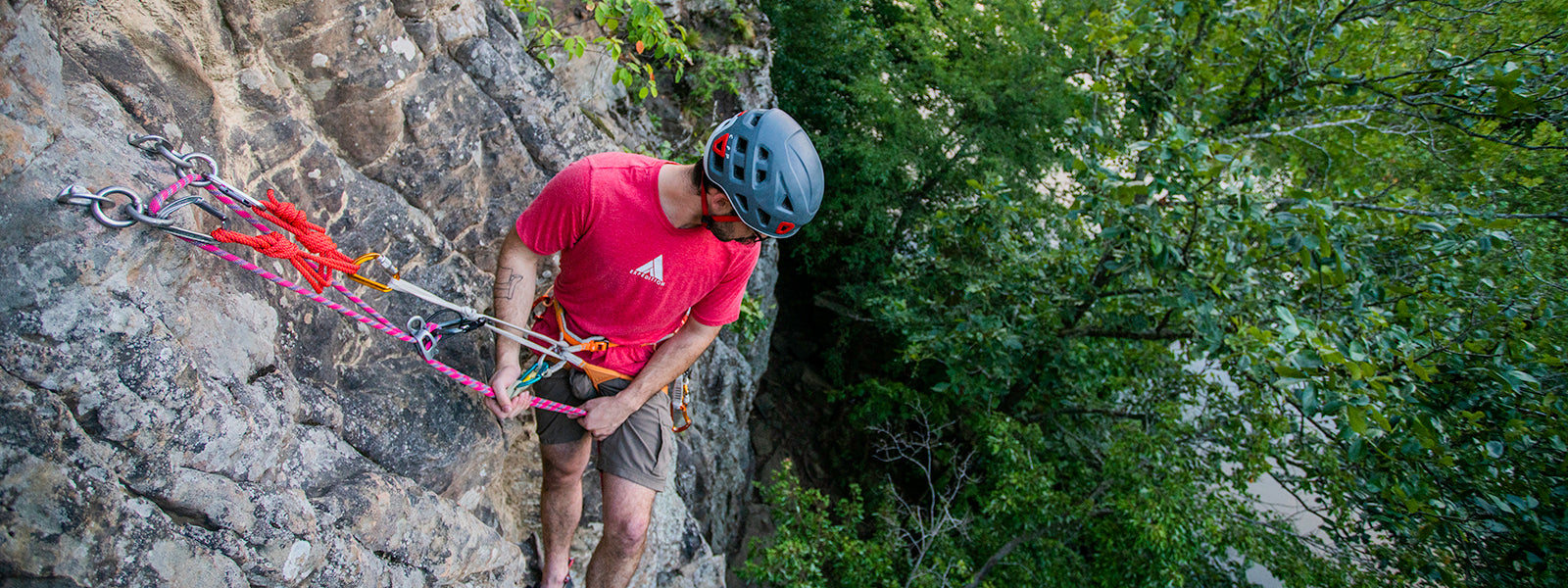 A man wearing a red shirt and climbing helmet looks at the ground from the top of a climbing route. He is about to rappel down the cliff.