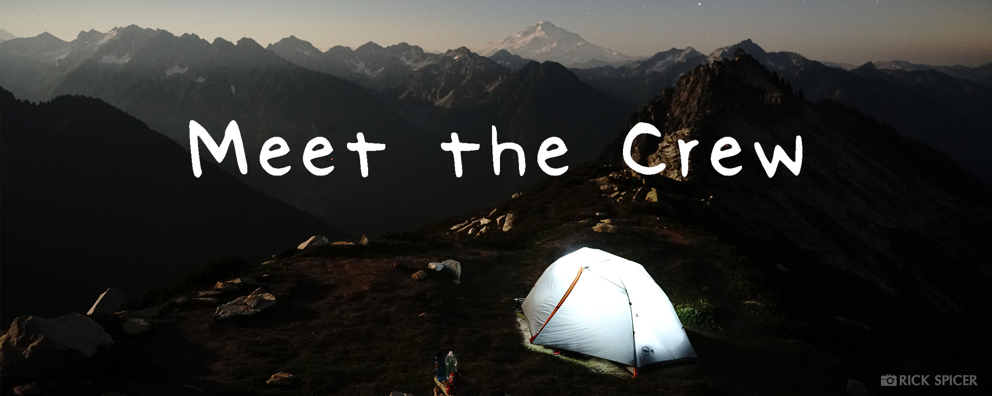 A tent set up in the North Cascades is lit up internally by a helmet, almost glowing. Mountains can be seen in the background.