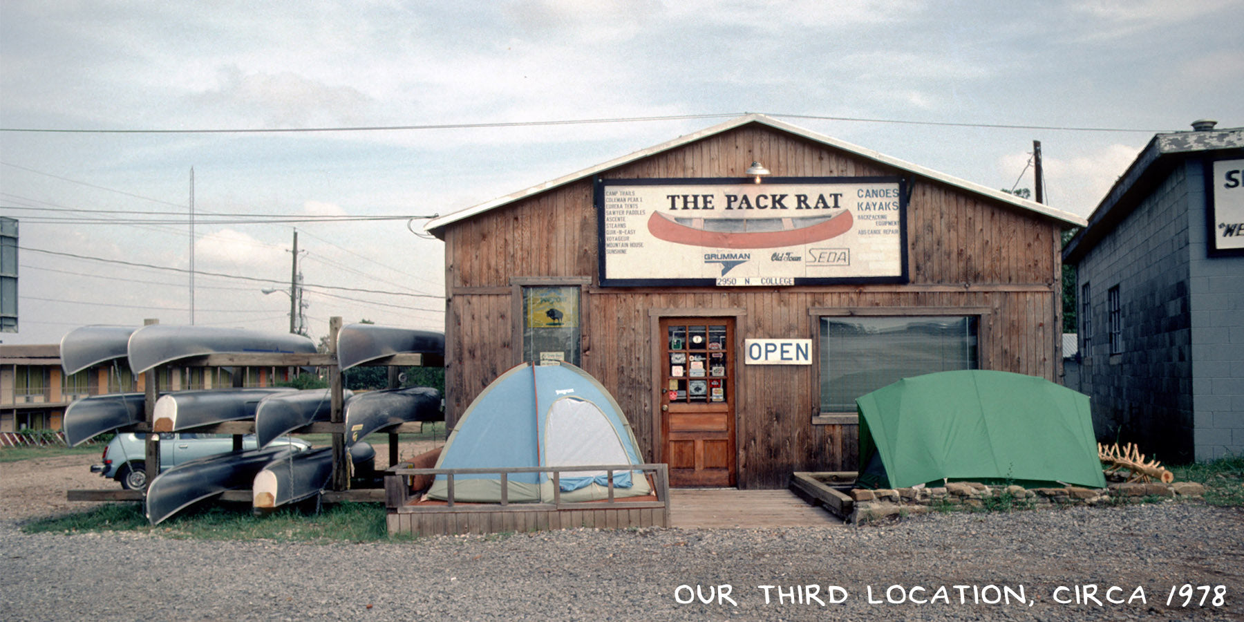 A wooden building with canoe and tents set up outside. The building's sign reads "The Pack Rat". An image of the third location of Pack Rat Outdoor Center.