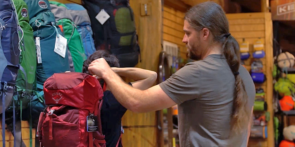 A man with a ponytail adjust a backpack to fit a customer. There is a wall of backpacks to their left.